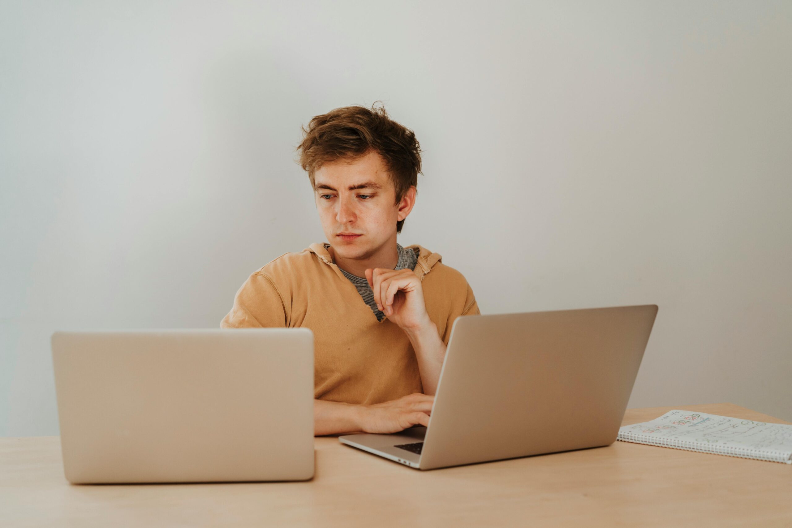 Focused young man working on two laptops in a home office setting, with a notebook visible on the desk.